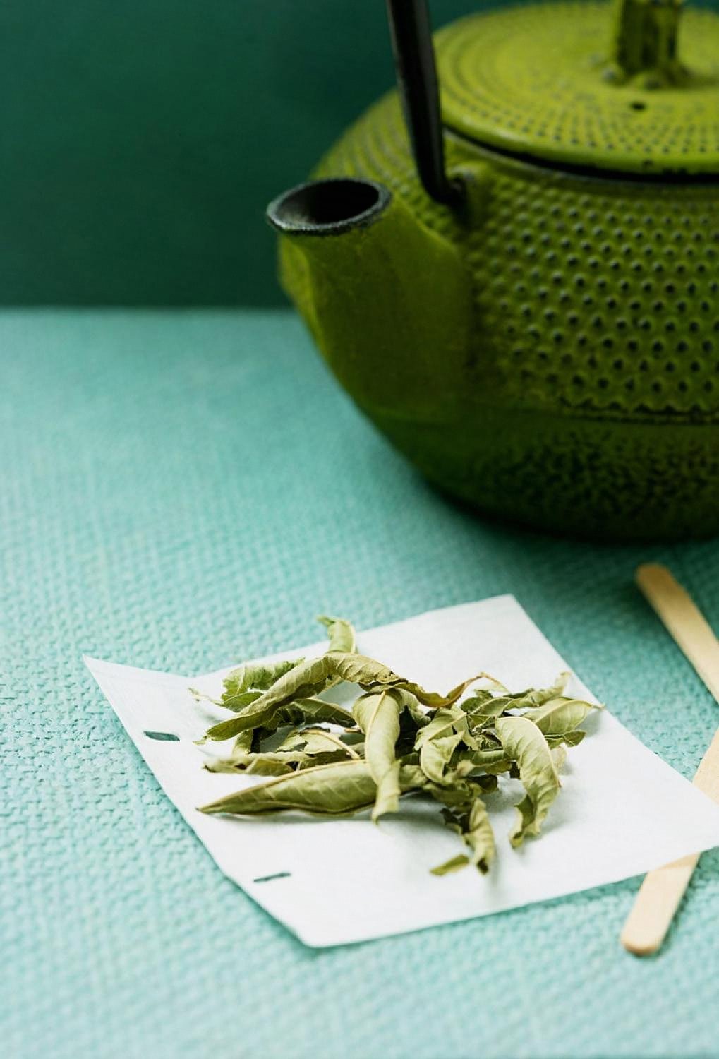 Dried verbena leaves on parchment beside a green cast-iron teapot